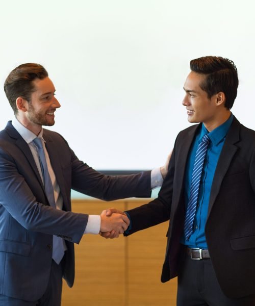 Closeup of two smiling middle-aged business men shaking hands and greeting each other in conference room with whiteboard in background